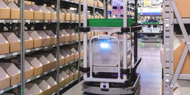 Autonomous robot navigating a warehouse aisle with shelves of cardboard boxes.