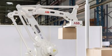 White robotic arm labeled ABB lifting a cardboard box in an industrial setting.