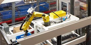 Yellow robotic arm operates in an automated warehouse with conveyor belts and storage bins.