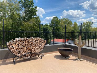 Outdoor patio with stacked firewood, a fire pit, and a cactus sculpture under a blue sky.
