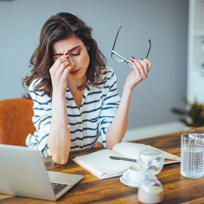 Tired woman holding glasses, rubbing her eyes at a desk.