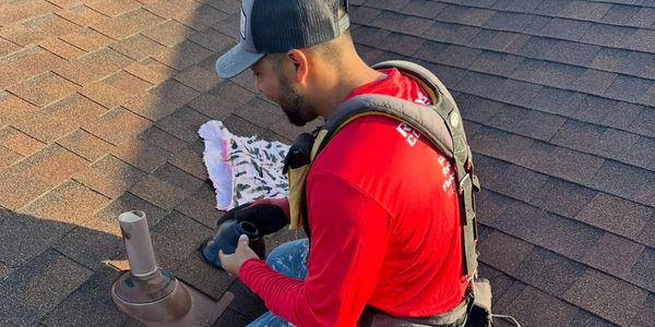 Roofer installing a vent pipe on a shingled roof with tools nearby.