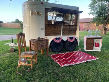 Patriotic outdoor setup with a vintage trailer, rustic furniture, and American flag decorations.