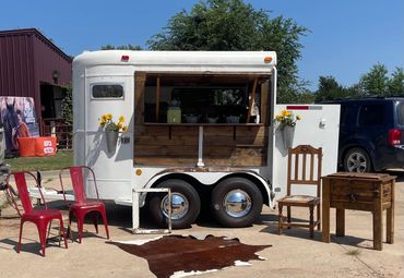 White trailer converted into a rustic outdoor bar with wooden accents and flowers.