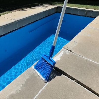 Blue pool brush on a telescopic pole resting on the stone coping beside clear water at a Prosper TX