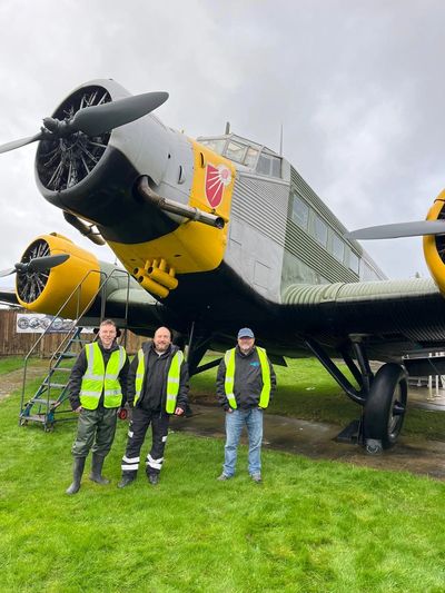 Three men in safety vests stand by a vintage aircraft.