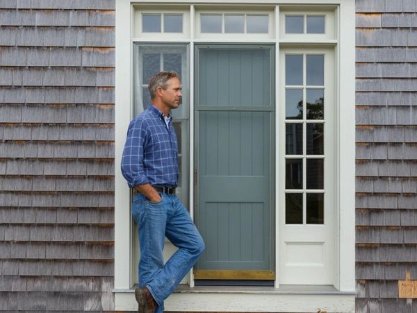 Man in blue shirt and jeans standing by a blue door of a house with wooden shingles.