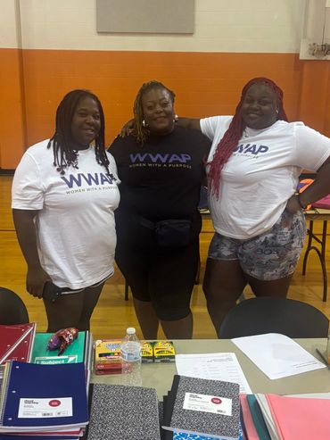 Three women wearing WWAP shirts stand behind a table with school supplies.