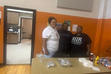 Three women smiling behind a table with food and condiments in a gymnasium.