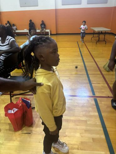 Young child in a yellow hoodie stands in a gymnasium with others seated and playing.