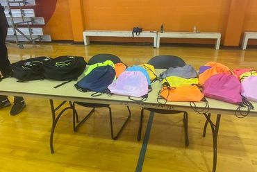Colorful drawstring bags and black backpacks arranged on a table in a gymnasium.