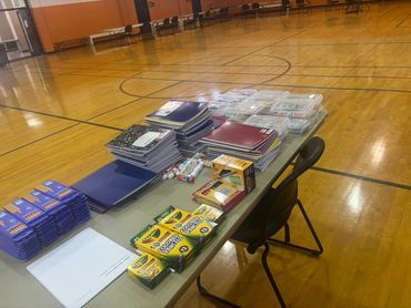 Table with school supplies like notebooks, colored pencils, and glue sticks in a gym.