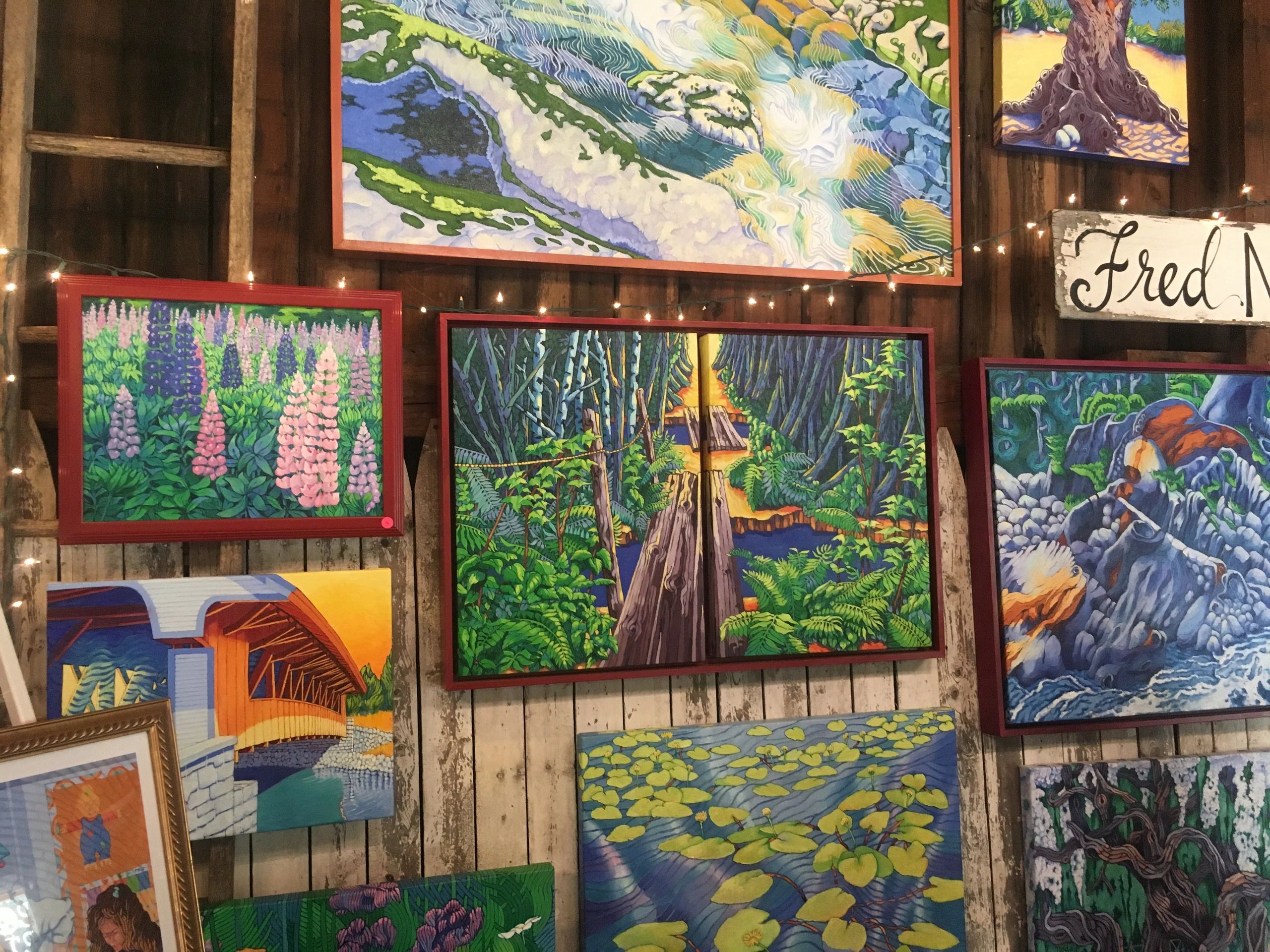 Colorful landscape paintings displayed on a rustic wooden wall with string lights.