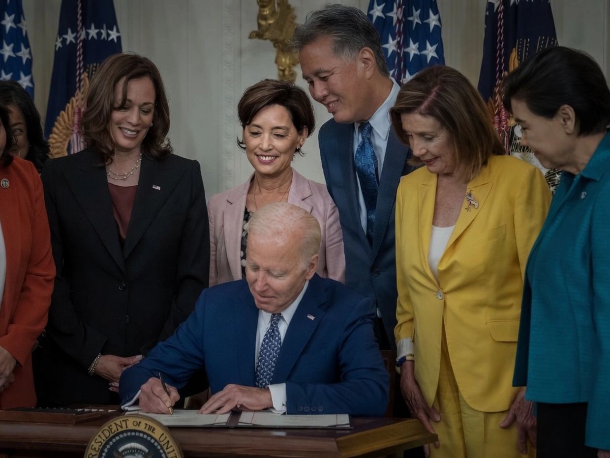 President Biden signs a document surrounded by smiling officials in a formal setting.