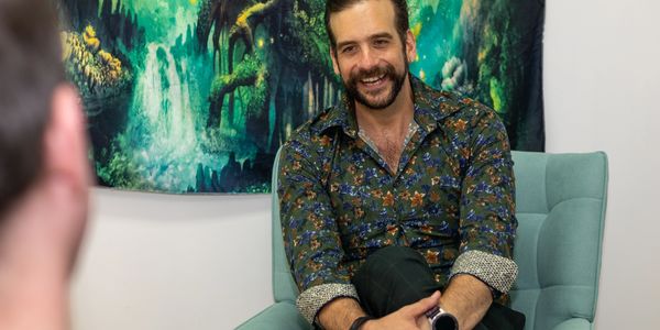 Man in floral shirt smiling and sitting cross-legged in a teal chair.