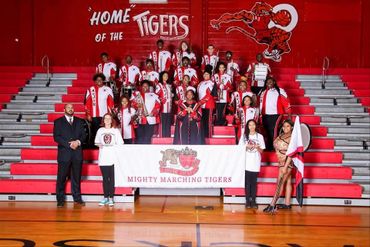 High school marching band posing in gym with 'Mighty Marching Tigers' banner.
