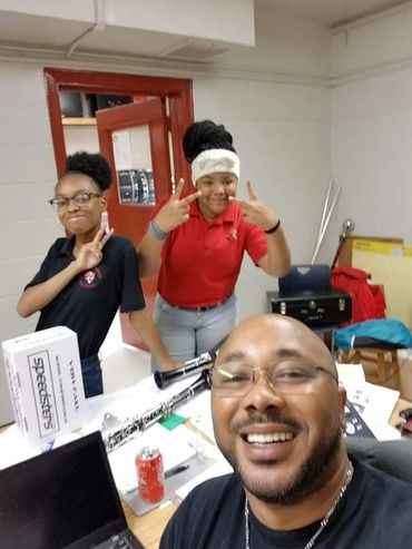 Three people smiling and posing for a selfie in a classroom with musical instruments.