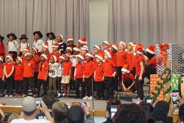 Children in red shirts and Santa hats perform on stage for a holiday event.