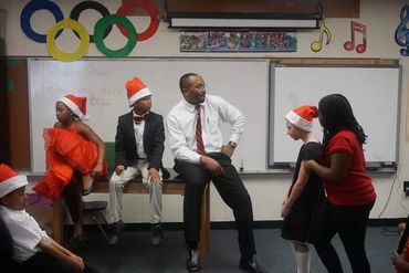 Children and a man in festive hats gather in a classroom decorated with Olympic rings and musical notes.