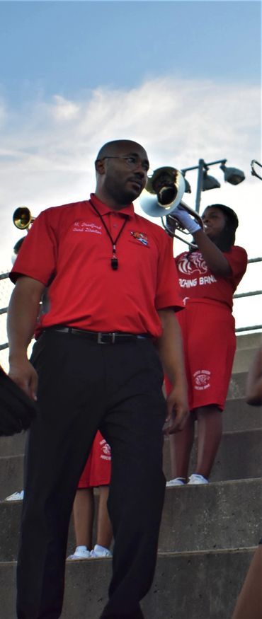 Band director standing in front of musicians in red uniforms playing brass instruments.