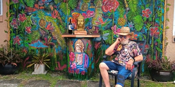 Man in colorful clothes sitting by vibrant, tropical-themed mural and sculpture.