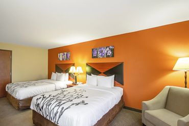 Hotel room with two beds, orange accent wall, and cozy armchair in Madison, WI. Accommodations for the Survive & Thrive 8791 Mental Health & Recovery Conference.
