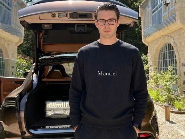 A man in glasses stands in front of an open car trunk with a suitcase inside.
