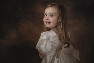 Little fairhaired girl in an ivory dress looking cheakily over her shoulder at the camera.