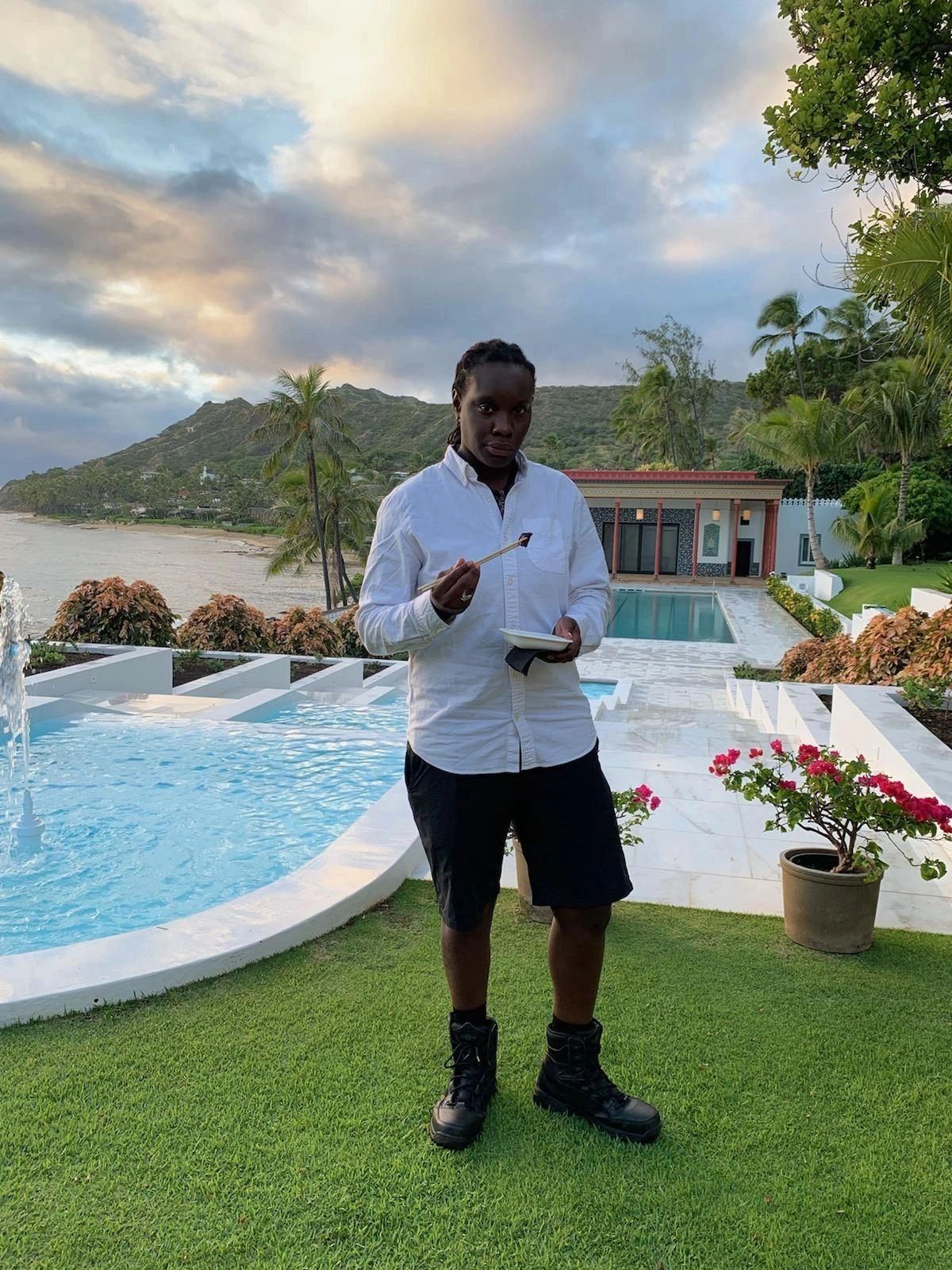 Person enjoying food near a pool with mountain and ocean views at sunset.