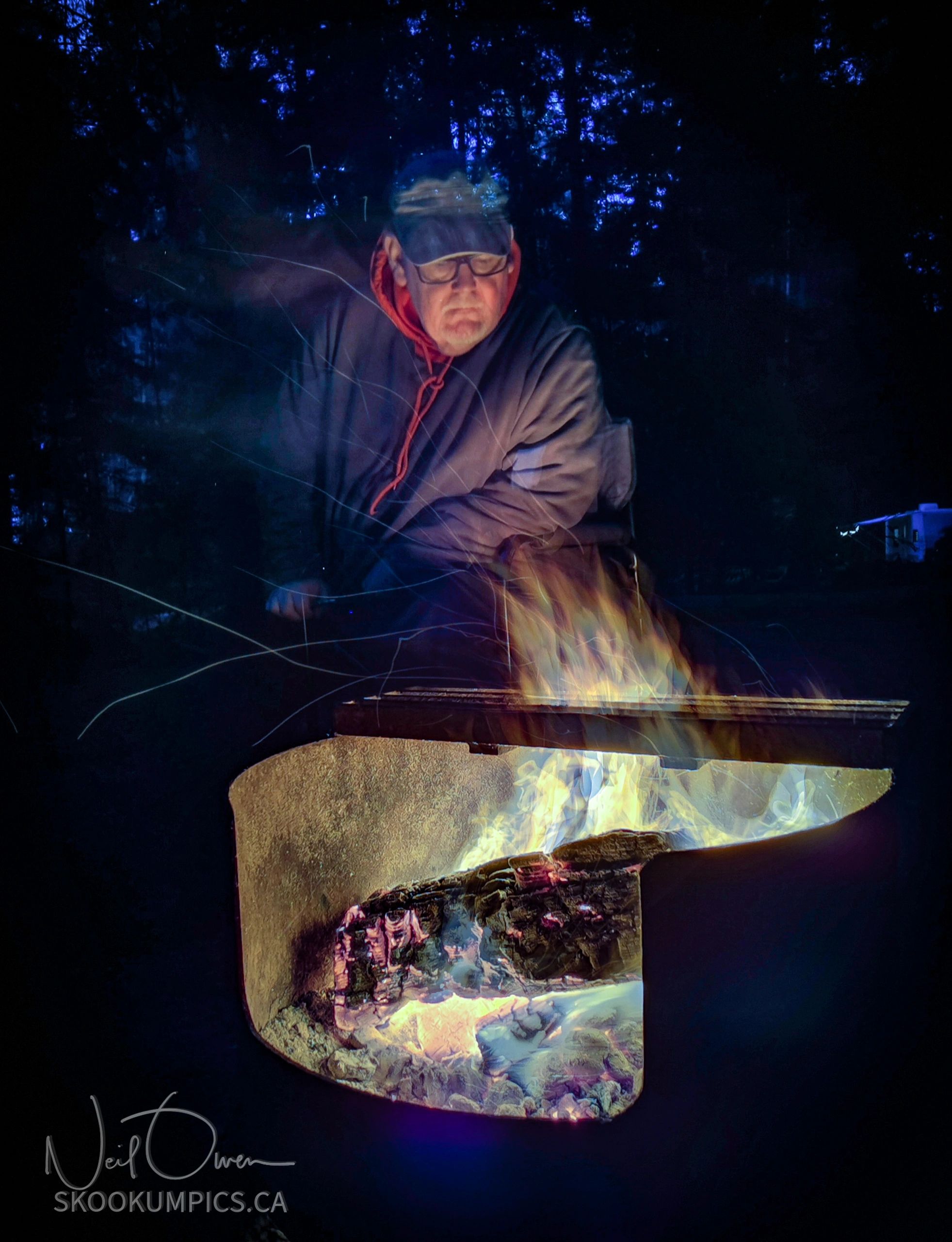 Man sitting beside fire in this long exposure image, Flames swirling as sparks dance in the smoke.