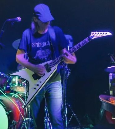 Guitarist playing a V-shaped electric guitar on stage with drums in the background.