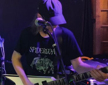 Musician playing guitar wearing a Spidergoat T-shirt and a cap on stage.