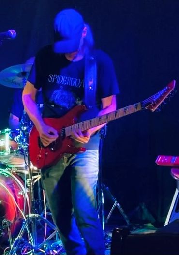 Musician playing electric guitar on stage with drum set in background.