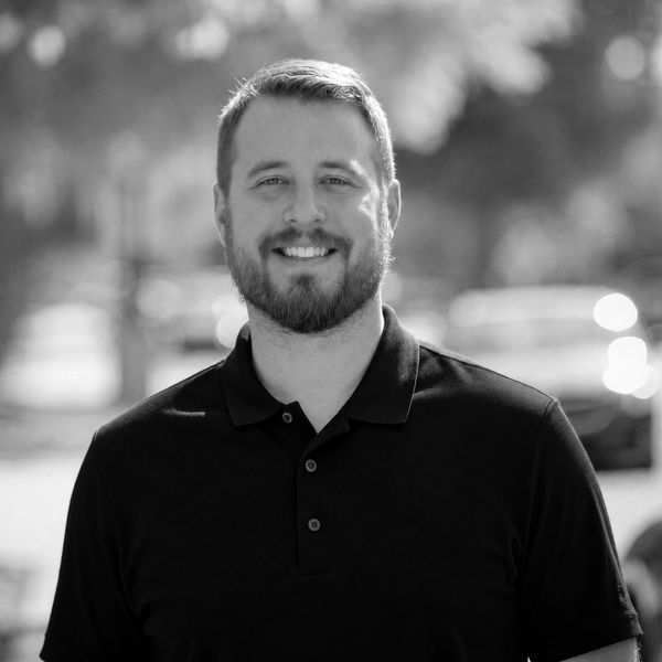 Smiling man with beard in a black polo shirt outdoors.
