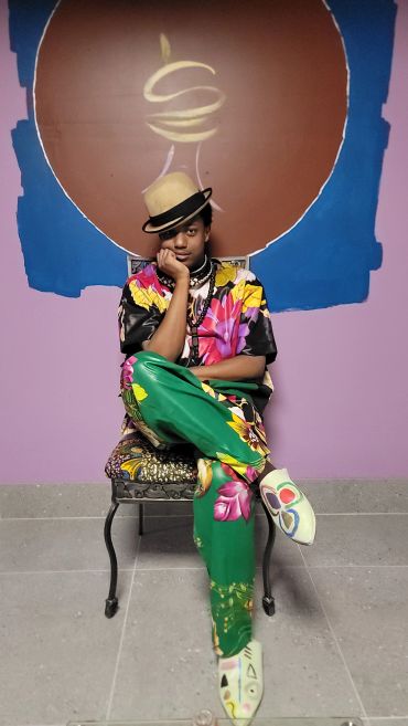 Person in vibrant floral outfit and hat sitting on a chair.