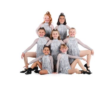 Seven young girls in silver fringe dance costumes posing in a pyramid formation.