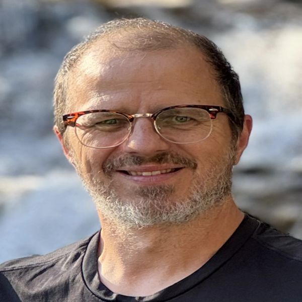 Smiling man with glasses and a beard outdoors.
