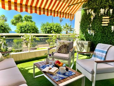Cozy balcony seating area with striped canopy and greenery under bright sunlight.