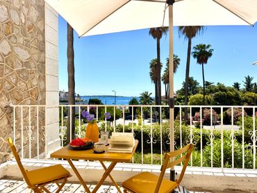 Sunny balcony with a table set for breakfast overlooking greenery and the sea.