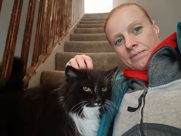 Person taking a selfie with a black and white cat on carpeted stairs.