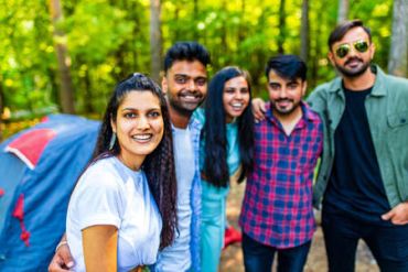 A group of five friends smiling outdoors near a tent in the forest.