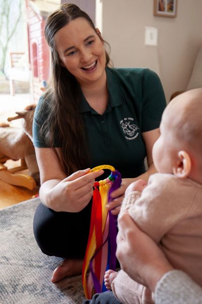 Woman with brown hair smiling at baby in mothers lap while playing with bright ribbon toy.