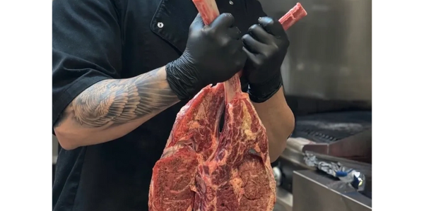 Chef proudly holding two large raw tomahawk steaks in a kitchen.