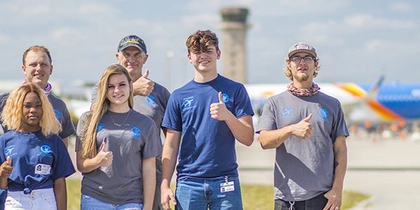 A group of young people and adults giving thumbs up outdoors near an airport runway.