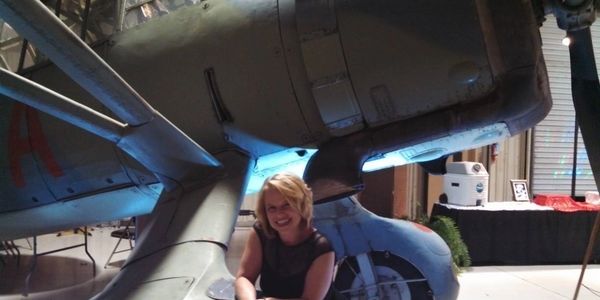 A woman smiling under the wing of a vintage aircraft indoors.