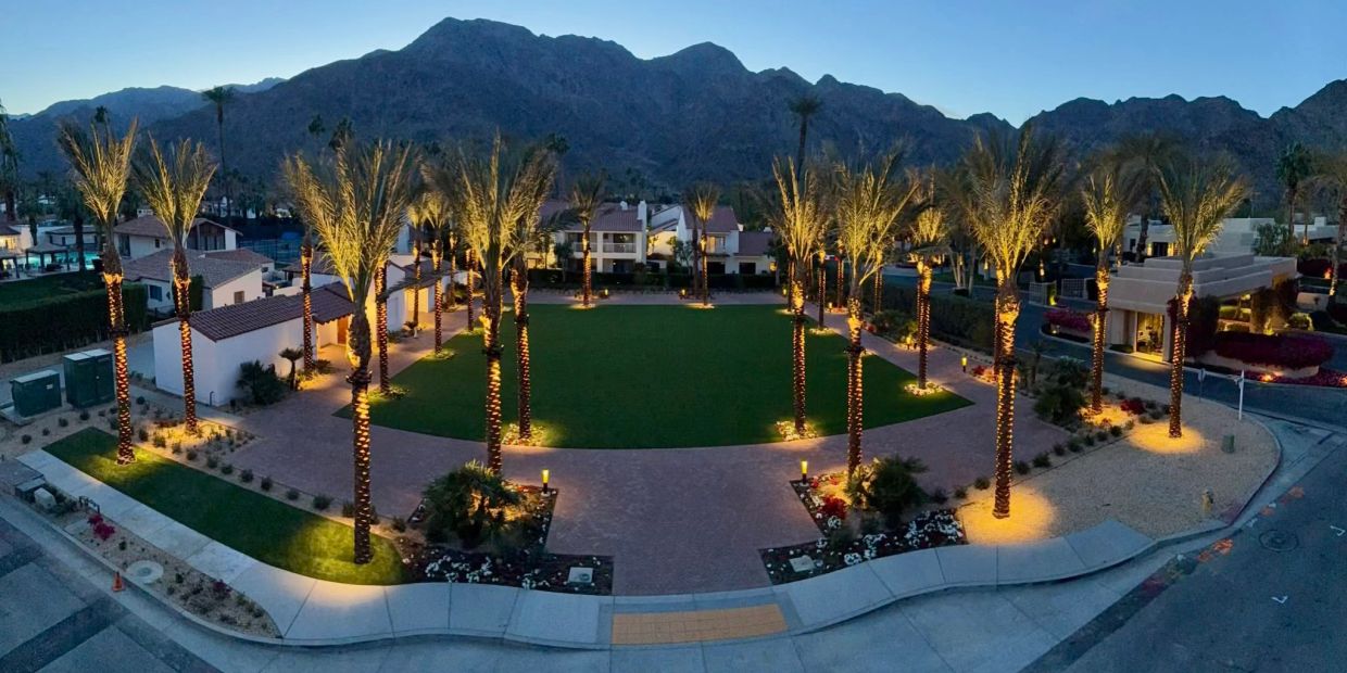 Palm trees lit at night around a square lawn with mountains in the background.