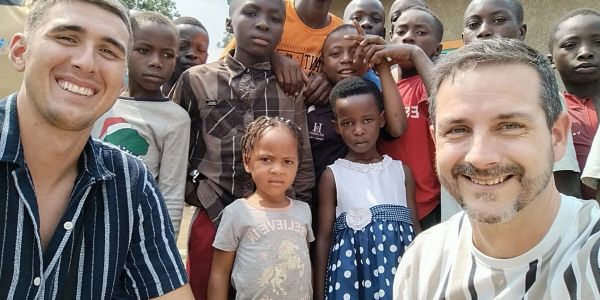 Zwei lächelnde Männer machen ein Selfie mit einer Gruppe afrikanischer Kinder.