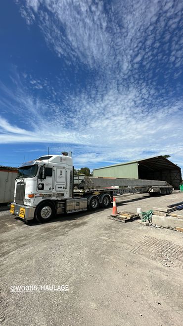 White heavy-duty truck hauling an oversized industrial beam under a partly cloudy sky.