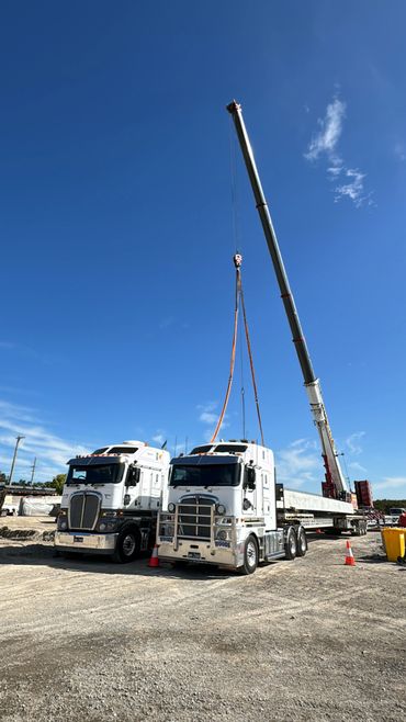 Two white trucks and a crane lifting a long load on a construction site.