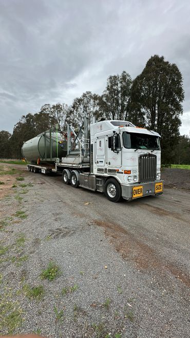 White Kenworth truck hauling a large green industrial tank on a flatbed trailer.